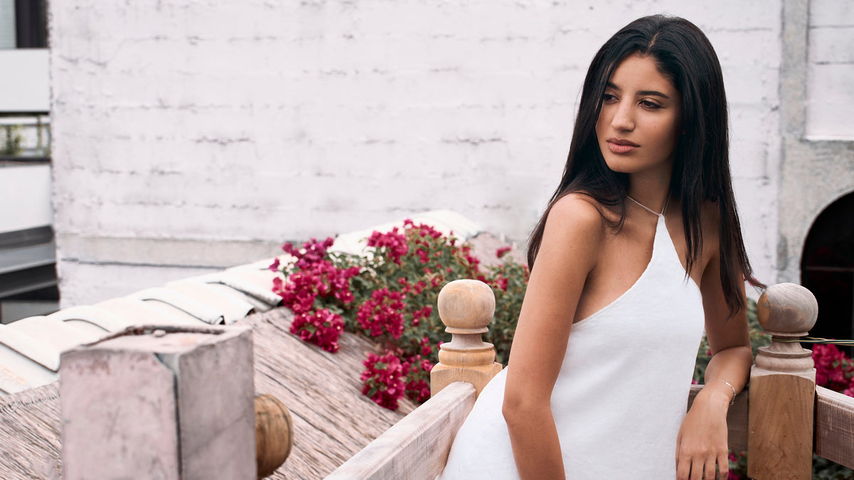 A woman with long dark hair wearing a white halter-neck dress, standing on a wooden balcony with white brick walls and red flowers in the background.
